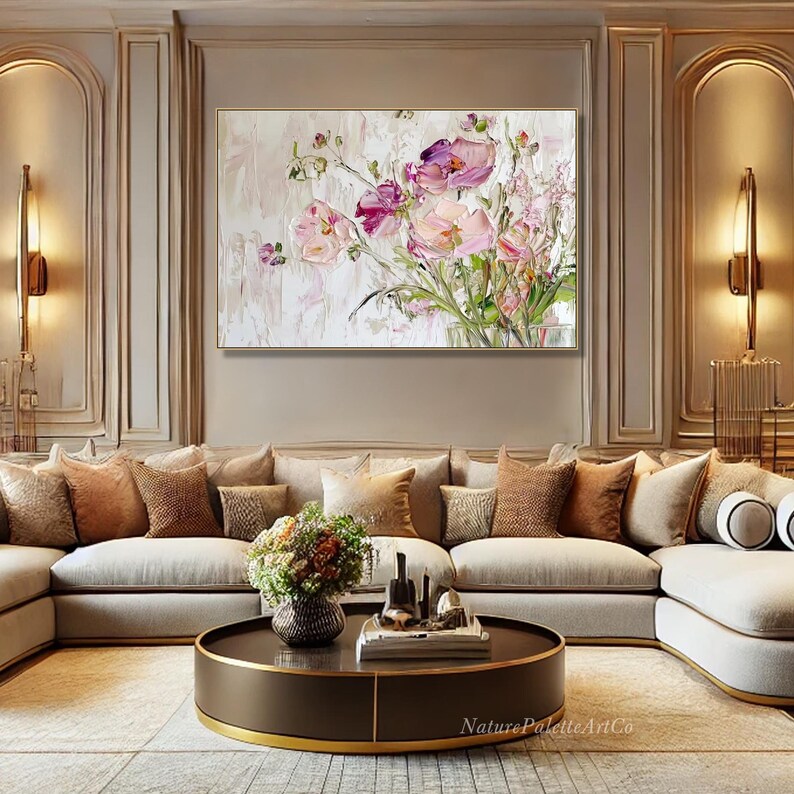 May include: A large, abstract painting of pink and purple flowers in a vase. The painting is on a white background and is framed in a gold frame. The painting is hanging on a wall in a living room with a beige sectional sofa and a brown coffee table.