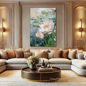 May include: An oil painting of a water lily with white and pink petals and yellow centers, surrounded by green lily pads. The artwork is displayed above a beige sectional sofa with decorative pillows. The painting is framed in black.