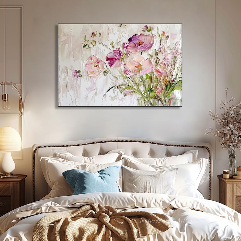 May include: A painting of a bouquet of pink and purple flowers with white accents on a white background. The painting is framed in black and is hanging above a bed with white bedding and pillows.