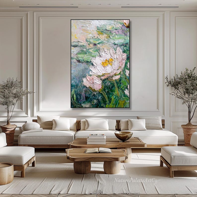 May include: Large framed oil painting featuring a close-up of white and pink water lilies with yellow centers, set against a backdrop of green lily pads and a soft, impressionistic background. The artwork is displayed in a modern living room.