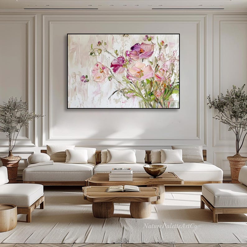 May include: A large abstract painting of pink and white flowers in a vase. The painting is hanging on a white wall in a living room setting with a white sofa and a wooden coffee table.