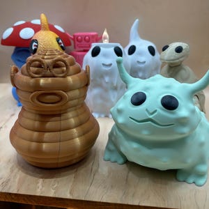Peut inclure: Une collection de figurines fantaisistes imprimées en 3D. L'assortiment comprend un champignon, un robot, une bougie et plusieurs créatures de différentes couleurs, dont le marron, le blanc et le bleu clair. La bougie est allumée avec une petite flamme.