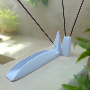 Könnte beinhalten: Ein hellblauer Räucherstäbchenhalter mit einer gebogenen Rinne und zwei vertikalen Spitzen. Drei schwarze Räucherstäbchen sind in den Halter gesteckt und geben Rauchschwaden ab. Der Halter steht auf einer hellen Oberfläche, mit grünem Laub im Hintergrund.