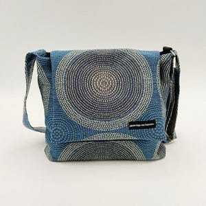 May include: A blue crossbody bag with a circular pattern design. The bag has a flap closure and an adjustable strap. The pattern is a series of concentric circles in shades of blue and off-white. The bag has a small black tag that reads "skinte bags, san francisco."