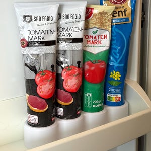 Könnte beinhalten: Im Kühlschrank befinden sich mehrere Tomatenmark-Tuben und eine blaue Soßen-Tube. Die Tomatenmark-Tuben sind weiß mit schwarzen und roten Etiketten. Die blaue Tube hat roten Text und eine gelbe Blume.