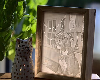 May include: A white ceramic cat figurine with gold spots, sitting next to a wooden picture frame with a black and white photo of a boxer dog inside. The photo is in a sepia tone and the dog is wearing a red bow tie.