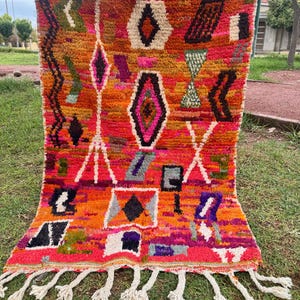 Peut inclure: Un tapis tissé à la main aux couleurs vives et au design géométrique. Le tapis présente un mélange de teintes orange, rouge, rose et violet, avec des franges blanches sur le bord inférieur. Le motif comprend des losanges, des triangles et des rectangles.