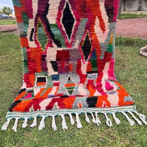 Peut inclure: Un tapis tissé à la main aux couleurs vives et au design géométrique. Le tapis présente un mélange de couleurs, notamment du rouge, de l'orange, du rose et du noir, avec des formes de losanges et rectangulaires. Il a une frange de pompons blancs le long du bord inférieur.