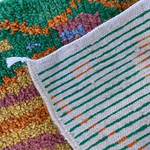 May include: Close-up of a colorful rug with a textured, woven design. The rug features a vibrant mix of green, orange, yellow, and purple hues. The reverse side displays a white background with green and orange stripes. A white tassel is visible.
