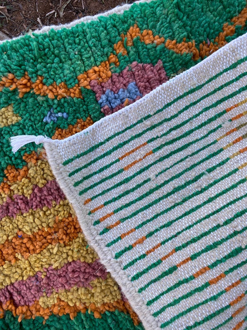 May include: Close-up of a colorful rug with a textured, woven design. The rug features a vibrant mix of green, orange, yellow, and purple hues. The reverse side displays a white background with green and orange stripes. A white tassel is visible.