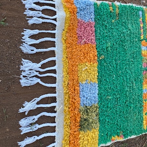 May include: A colorful, rectangular rug with a textured, woven design. The rug features a vibrant mix of colors, including green, orange, yellow, blue, and pink. White fringe adorns one edge. This rug is suitable for adding a pop of color to any room.