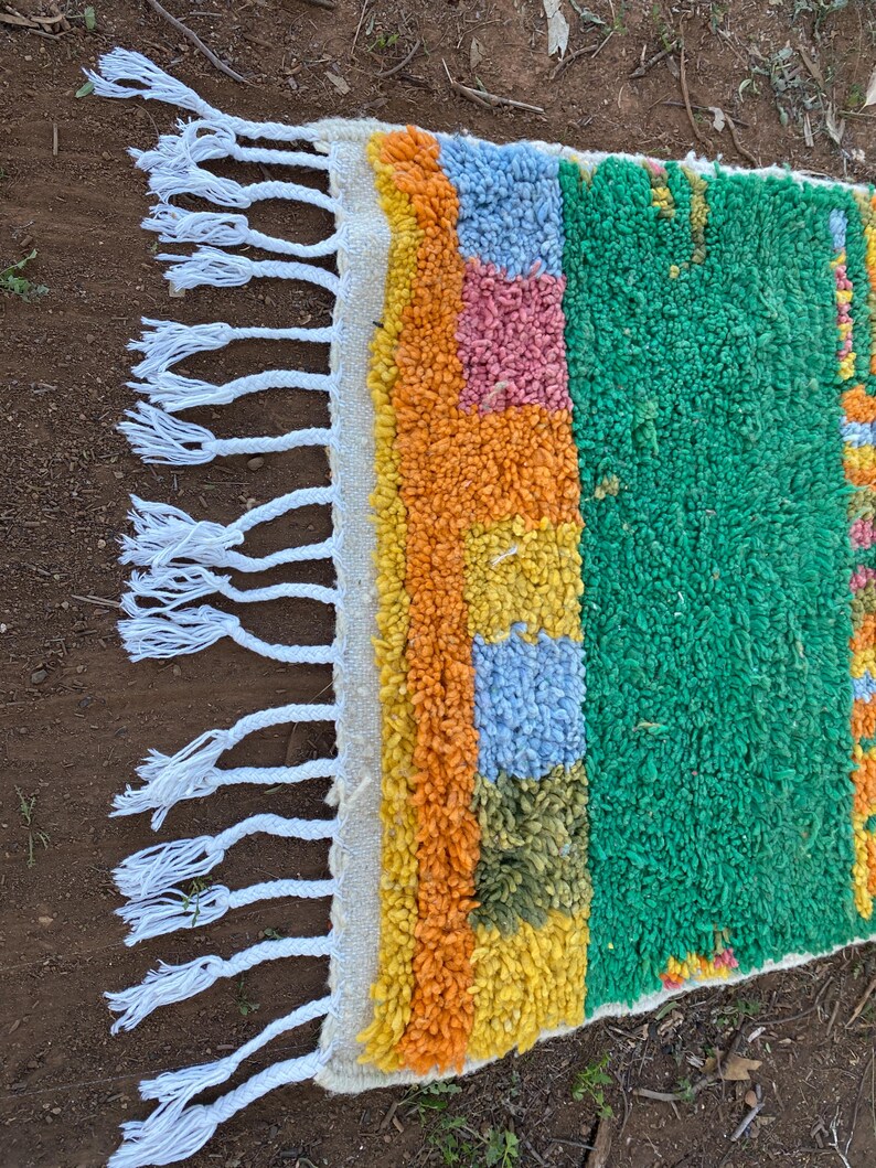 May include: A colorful, rectangular rug with a textured, woven design. The rug features a vibrant mix of colors, including green, orange, yellow, blue, and pink. White fringe adorns one edge. This rug is suitable for adding a pop of color to any room.