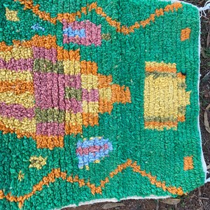 May include: A hand-woven rug with a vibrant green background and colorful geometric patterns. The design incorporates shades of orange, yellow, pink, and blue, creating a textured and visually appealing piece. The rug's edges are finished with a white border.