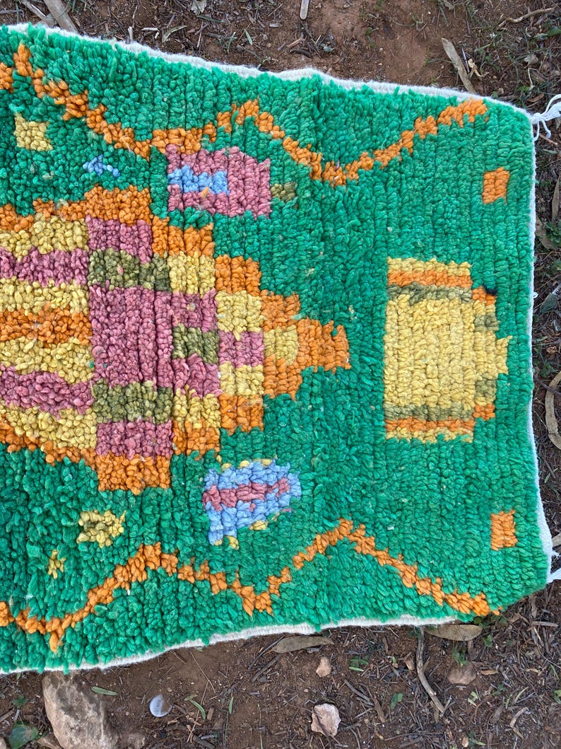 May include: A hand-woven rug with a vibrant green background and colorful geometric patterns. The design incorporates shades of orange, yellow, pink, and blue, creating a textured and visually appealing piece. The rug's edges are finished with a white border.