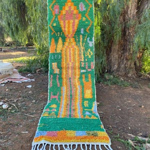 May include: A vibrant, rectangular rug with a green background and colorful geometric patterns in orange, yellow, pink, and blue. The rug has a fringed edge and is displayed outdoors, showcasing its textured design and bright colors.