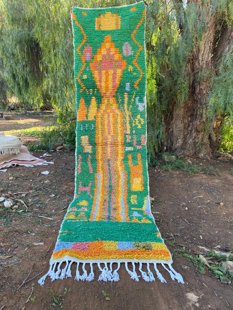 May include: A vibrant, rectangular rug with a green background and colorful geometric patterns in orange, yellow, pink, and blue. The rug has a fringed edge and is displayed outdoors, showcasing its textured design and bright colors.