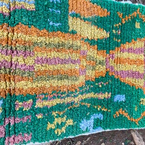 May include: A colorful, textured rug with a vibrant design. The rug features a mix of green, orange, yellow, pink, and blue hues. The rug's design includes geometric and abstract patterns, creating a visually appealing textile.