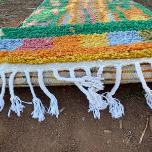 May include: A colorful, handmade rug with a textured surface. The rug features a vibrant mix of colors, including green, orange, yellow, pink, and blue. White tassels hang from the edge. This decorative rug is perfect for adding a touch of bohemian style to any room.