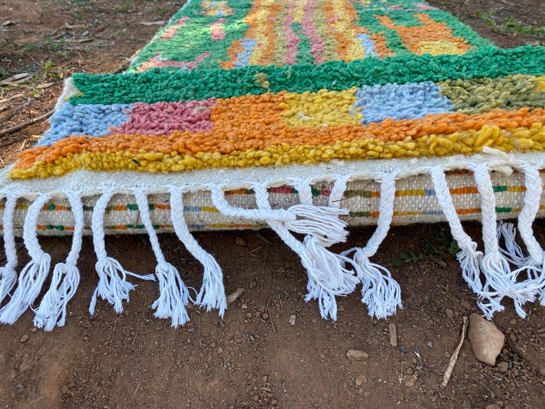 May include: A colorful, handmade rug with a textured surface. The rug features a vibrant mix of colors, including green, orange, yellow, pink, and blue. White tassels hang from the edge. This decorative rug is perfect for adding a touch of bohemian style to any room.