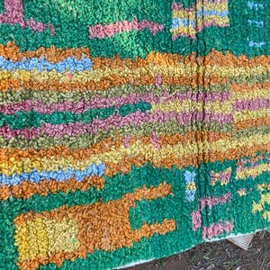 May include: A colorful, textured rug with a green background and a pattern of orange, yellow, pink, and blue shapes. The rug has a thick, plush pile, and the design is abstract and geometric.