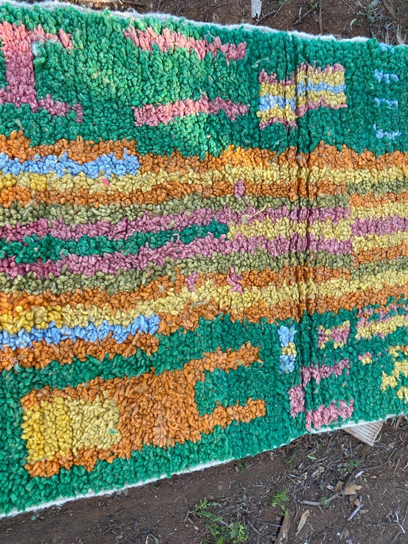 May include: A colorful, textured rug with a green background and a pattern of orange, yellow, pink, and blue shapes. The rug has a thick, plush pile, and the design is abstract and geometric.