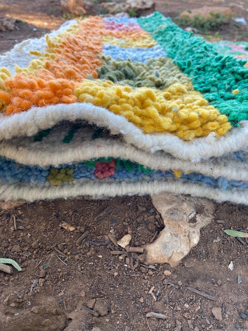 May include: A colorful, handmade rug with a geometric design. The rug features a variety of colors, including orange, yellow, green, and blue. The rug is made of a thick, textured material, and the edges are finished with a white border. The rug is folded over.