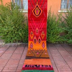 Peut inclure: Un tapis long et étroit avec un motif géométrique coloré. Le tapis est principalement rouge, orange et rose, avec des accents noirs et verts. Il a des franges aux deux extrémités.