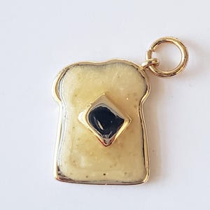 Könnte beinhalten: Goldfarbener Anhänger in Form einer Toastscheibe mit gelb-cremefarbener Oberfläche. Ein dunkelblauer, rautenförmiger Akzent befindet sich in der Mitte des Toasts. Der Anhänger hat einen goldenen Rand und eine kleine Schlaufe für eine Kette.