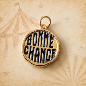 May include: A round gold-toned metal charm featuring a scalloped edge and a small jump ring at the top. The center is filled with dark blue enamel and displays the text BONNE CHANCE in raised gold-toned capital letters across two lines.