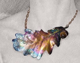 May include: A necklace featuring a large, iridescent oak leaf pendant with vibrant purple, blue, and gold tones, attached to a dark metal chain. The pendant includes a small, round, brown bead resembling an acorn attached to the leaf stem.