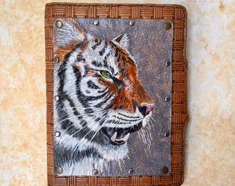 May include: A brown leather journal with a woven border and a painted tiger portrait. The tiger has orange and white fur, green eyes, and is set against a textured background. Metal rivets secure the portrait to the journal cover.