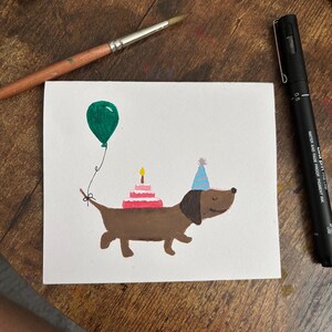 May include: A hand-painted birthday card showing a brown dachshund with a party hat and cake. A green balloon floats above the dog. A paintbrush and pen are on a wooden surface.