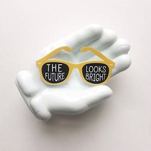 May include: A white ceramic hand holding a pair of yellow sunglasses with the text "THE FUTURE LOOKS BRIGHT" printed in black.