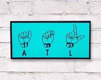 Könnte beinhalten: Ein türkisfarbenes Schild mit schwarzer Beschriftung und drei schwarzen Handzeichen, die das Wort "ATL" buchstabieren.
