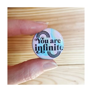 May include: A round button with a pastel pink, blue and white background. The button has black text that reads "You are infinite" with a stylised rainbow design.