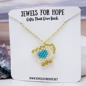 Peut inclure: Collier en chaîne dorée avec un pendentif en perles blanches et turquoise en forme de tasse à thé. Le collier est présenté sur une carte blanche avec le texte "Jewels for Hope Gifts That Give Back" et "www.jewelsforhope.net".