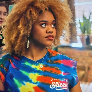 Peut inclure: Une femme aux cheveux blonds bouclés portant un t-shirt bleu tie-dye avec le texte "Salt Lake Slices" imprimé sur le devant.
