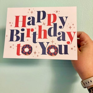 May include: A white card with the text "Happy Birthday to You" in red and blue lettering. The card has a glittery background and two donut illustrations.