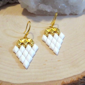 May include: A pair of gold-toned drop earrings with a geometric design. The earrings feature a diamond-shaped pattern of white beads, topped with a gold-colored, textured section. The earrings hang from gold-colored ear wires.