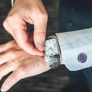 May include: A silver watch with a white face and Roman numeral hour markers. The watch has a square face and a metal band. The watch is on a person's wrist. The person is wearing a white dress shirt with cufflinks.