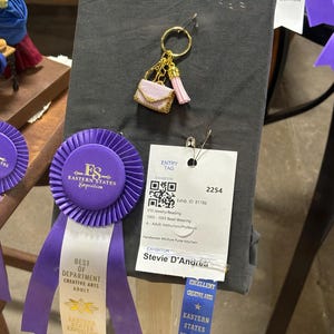 May include: A handwoven miniature purse keychain with a gold-colored ring, a pink tassel, and a small, beaded purse. The keychain is displayed on a gray fabric with award ribbons and an exhibitor tag that reads "Stevie D'Andrea."