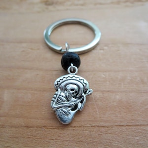 May include: Silver skeleton playing a guitar keychain with a black lava rock bead and a silver key ring.