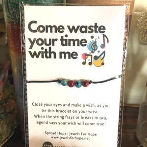 May include: A black string bracelet with red, blue, and white beads. The bracelet is tied around a white card with the text "Come waste your time with me" and musical notes. The card also includes the text "Spread Hope | Jewels For Hope" and "www.jewelsforhope.net".