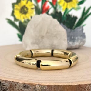 May include: A gold-toned bracelet composed of smooth, cylindrical metal beads alternating with small, thin black spacers. The bracelet features a polished, reflective finish and is arranged in a circular shape on a light-colored wooden surface.