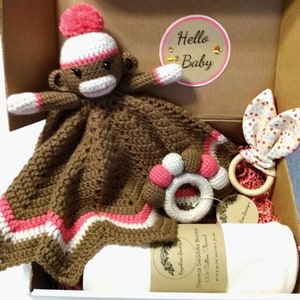 May include: A gift box filled with baby items, including a brown crocheted monkey toy, a brown and white crocheted blanket with pink trim, a wooden teething ring with a fabric bunny attached, and a white swaddle blanket. The box has a label that says "Hello Baby".