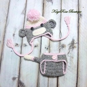 May include: A gray and pink crocheted monkey hat and diaper set for a baby. The hat has a pink pom pom on top and a long tail that ties around the neck. The diaper has a gray button in the front.