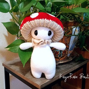 May include: A handmade crochet mushroom figure with a red cap and white spots, a beige frill, and a white body. The figure has large black eyes and small arms and feet. The mushroom is displayed on a wooden surface in front of a potted plant.