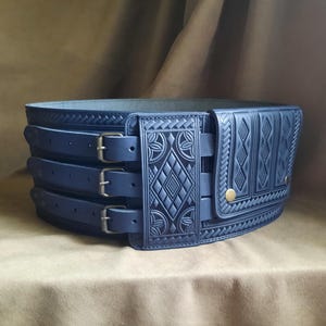 Op de afbeelding: Een donkerblauwe leren riem met ingewikkelde geometrische en bloemmotieven. De riem heeft drie verstelbare riemen met zilveren gespen en een decoratief zakje met een messing knoopsluiting. Handgemaakt ontwerp.