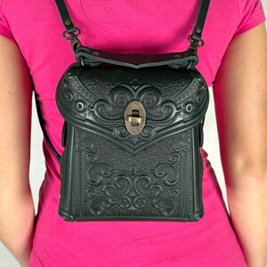 Pode incluir: Mochila de couro verde escuro com desenhos florais e de volutas em relevo. A mochila tem alças ajustáveis e um fecho de metal. A mochila é usada nas costas.