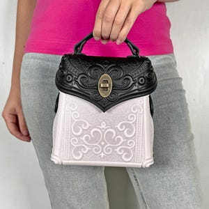 Könnte beinhalten: Eine kleine, verzierte Handtasche mit schwarzem Oberteil und Griff sowie einem weißen Körper mit Wirbelmuster. Die Tasche hat einen goldfarbenen Verschluss und Zierleisten.